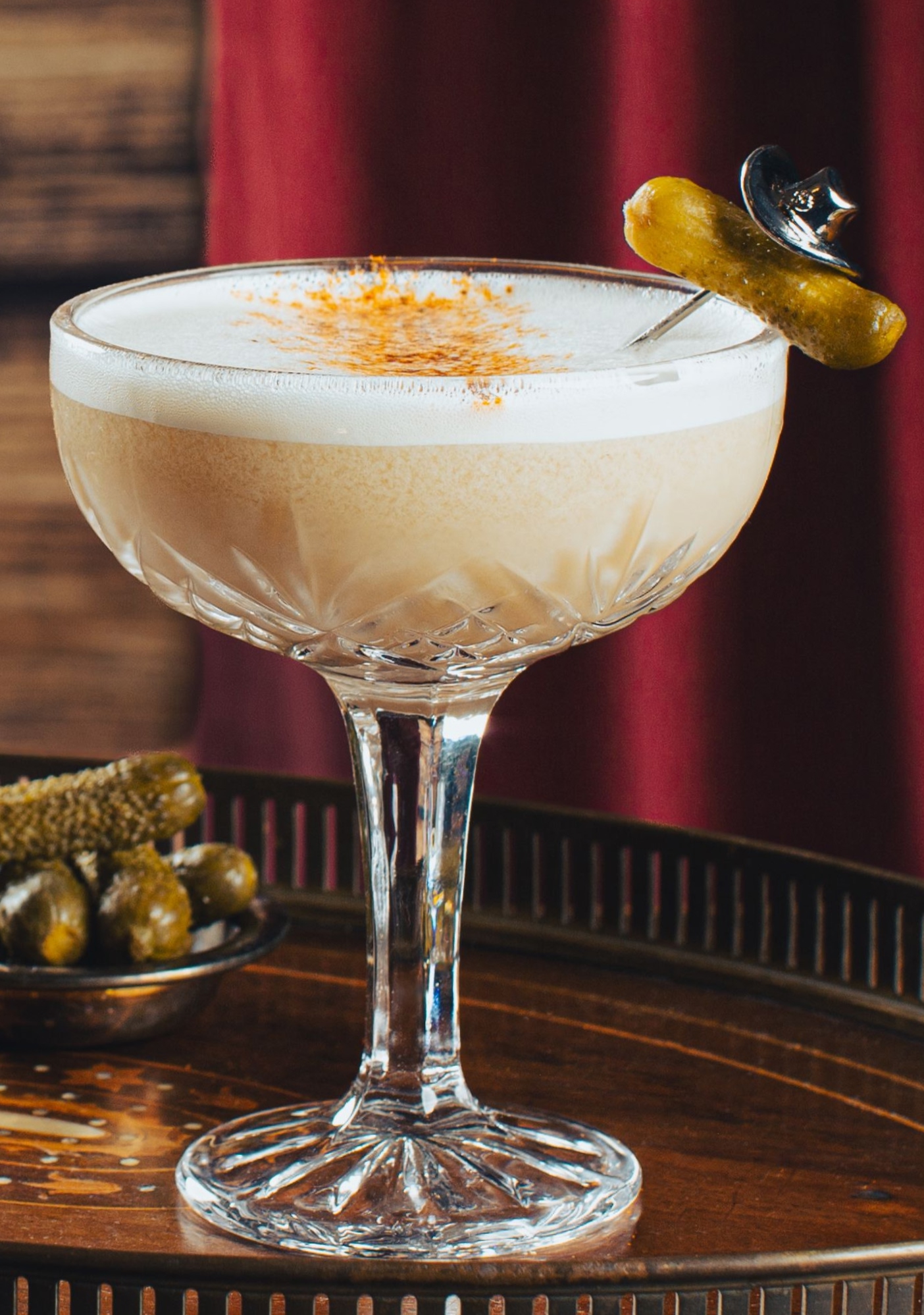 A cocktail in a coupe glass with a frothy top and a pickle garnish.