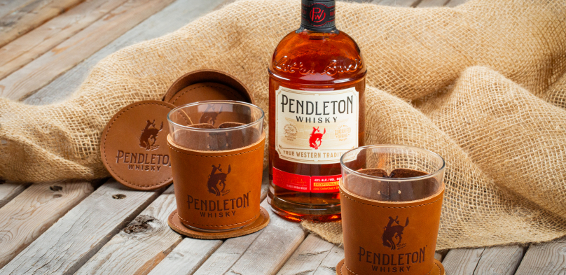 Bottle of Pendleton Whisky placed on burlap fabric alongside two branded whiskey glasses and leather coasters, displayed on a rustic wooden surface.