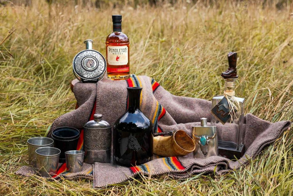 Antique Western flasks, cups, a horn and decanters arranged on a striped wool blanket with a bottle of Pendleton Whisky